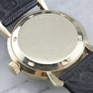 Pu&ograve; includere: Il retro di un orologio da polso color oro con cinturino in pelle nera. La cassa dell'orologio &egrave; incisa con "14 Karat Gold".