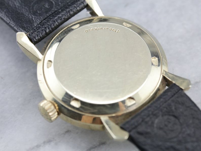 Pu&ograve; includere: Il retro di un orologio da polso color oro con cinturino in pelle nera. La cassa dell'orologio &egrave; incisa con "14 Karat Gold".