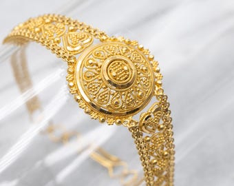 May include: A gold-colored bracelet with intricate filigree and a central circular design. The bracelet features detailed patterns and a chain-link band. The ornate jewelry is displayed against a soft, blurred background.