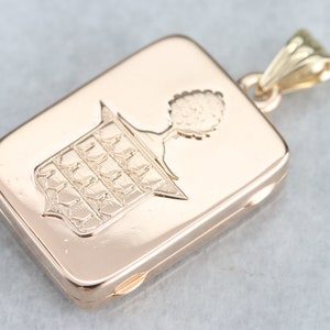 May include: A gold locket featuring an engraved design of a building with a spire on top. The locket has a hinged lid and a loop for attaching a chain.