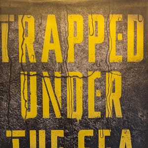 May include: A book cover with a black, textured background featuring yellow, distressed lettering that reads TRAPPED UNDER THE SEA. Below the title, white text reads ONE ENGINEERING MARVEL, FIVE MEN, AND A DISASTER TEN MILES INTO THE DARKNESS, followed by the author name NEIL SWIDEY.