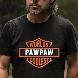 May include: Black t-shirt with an orange and white graphic that reads "Worlds Pawpaw Coolest". The design is in a shield shape. The person in the image is wearing the shirt outdoors.