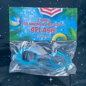 May include: A blue and white thank you card with the text "Thanks for making my birthday a splash! Love, Talia". The card is in a clear plastic bag with a pair of blue swimming goggles.