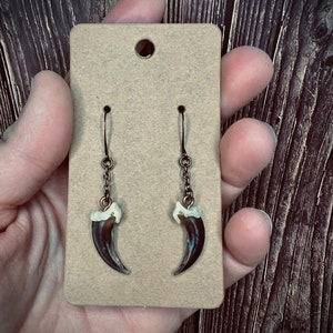 May include: A pair of dangle earrings featuring dark brown claw pendants with off-white accents. The earrings hang from bronze-colored hooks and chains, displayed on a tan earring card. The background is a dark wood grain.