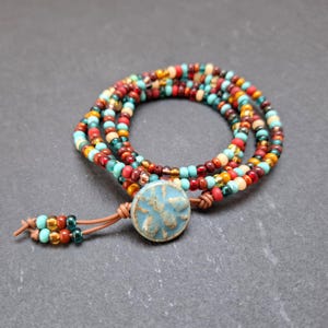 May include: A multi-strand beaded wrap bracelet. The bracelet features a mix of colourful beads in red, turquoise, gold, and brown. A light blue ceramic button with a starburst design serves as the clasp, and the bracelet is secured with a leather cord.