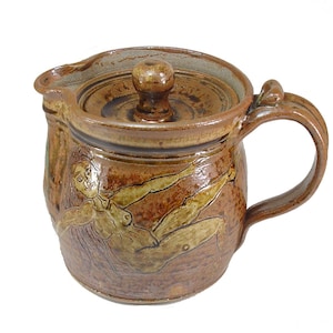 Peut inclure: Une théière en céramique brune avec un couvercle et une anse. La théière est recouverte d'un glaçage brun avec un motif de figure nue sur le côté.