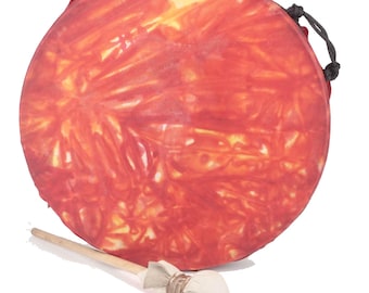 Puede incluir: Un tambor redondo de mano con un vibrante diseño tie-dye rojo y naranja. Una baqueta de madera con una cabeza de tela blanca descansa junto al tambor. Una cuerda negra está unida al tambor.