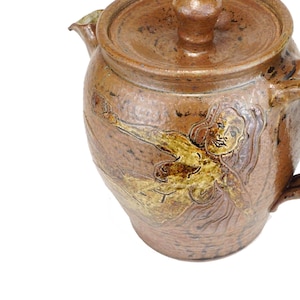 May include: A brown ceramic teapot with a lid and a handle. The teapot is decorated with a brown and gold image of a woman.