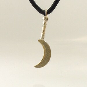 May include: A silver-colored pendant shaped like a small scythe, suspended from a black cord. The pendant has a curved blade and a textured handle, suggesting a miniature weapon or tool. The background is a plain, light color.