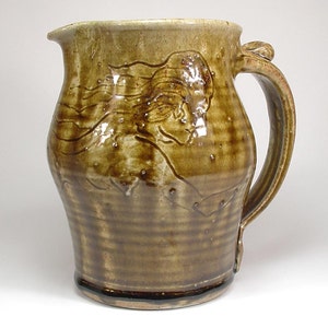 May include: A brown ceramic pitcher with a raised design of a woman's face and flowing hair. The pitcher has a wide base and a handle.