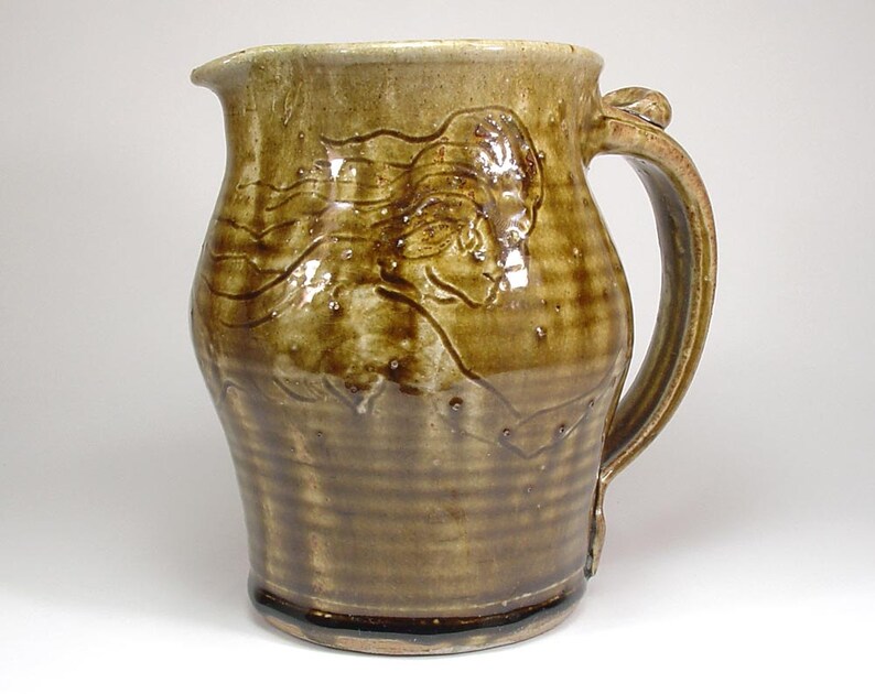 May include: A brown ceramic pitcher with a raised design of a woman's face and flowing hair. The pitcher has a wide base and a handle.