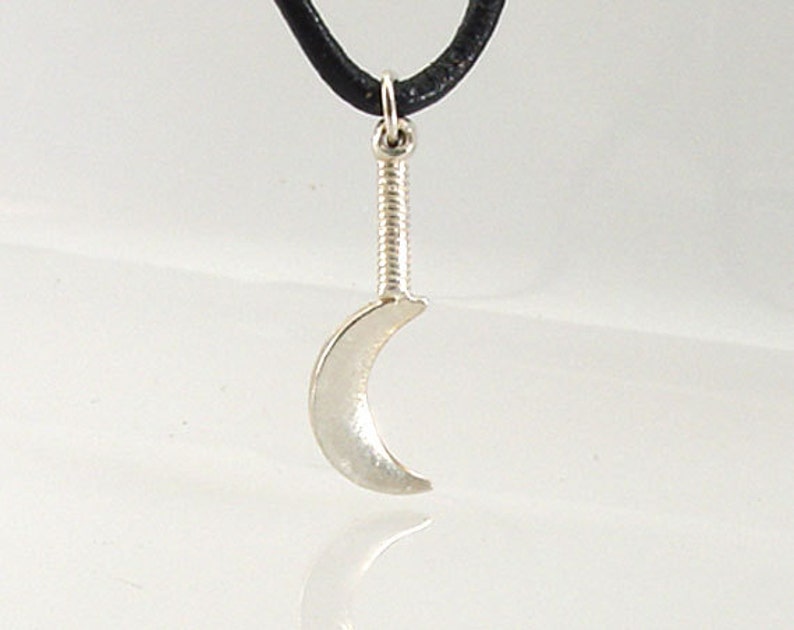 May include: A silver-colored crescent moon pendant with a textured handle, suspended from a black cord. The pendant is a stylized representation of a crescent moon. The design is simple and elegant, suitable for everyday wear.