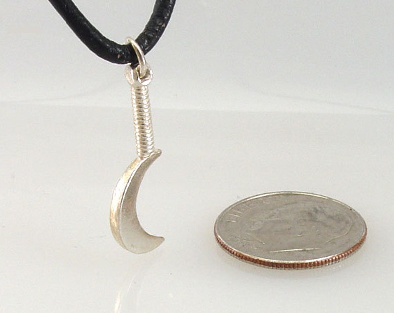 May include: A silver-colored pendant necklace featuring a small, curved blade design, suspended from a black cord. The pendant has a detailed handle and a crescent-shaped blade. A US quarter coin is next to the pendant for size comparison.