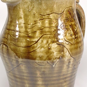May include: A brown ceramic pitcher with a textured surface and a carved design. The pitcher has a handle and a spout.