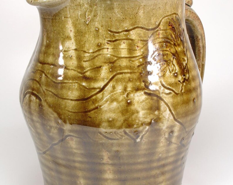 May include: A brown ceramic pitcher with a textured surface and a carved design. The pitcher has a handle and a spout.
