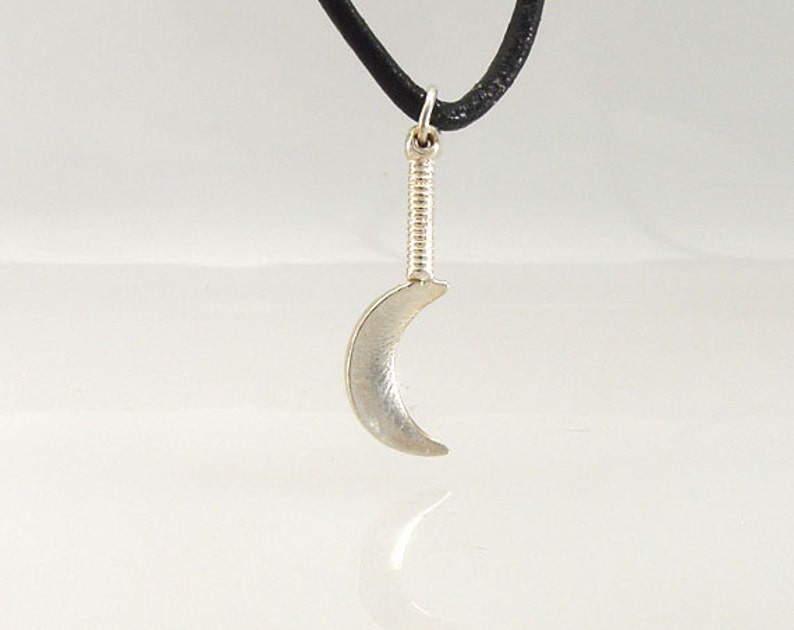 May include: A silver crescent moon pendant with a textured handle, suspended from a black cord. The pendant is a stylized representation of a crescent moon, with a smooth, curved surface. The handle has a ribbed design.