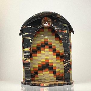 May include: A small, arched structure featuring a black exterior with swirling orange and yellow marbled patterns. The interior back wall displays a grid of small, rectangular tiles in shades of yellow, orange, brown, and black, creating a mosaic effect.