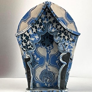 Peut inclure: Une structure en céramique avec un toit pointu et une ouverture arquée, présentant un motif marbré dans des tons de bleu, blanc et noir. L'extérieur affiche un design géométrique, tandis que les parois intérieures montrent un motif fluide.