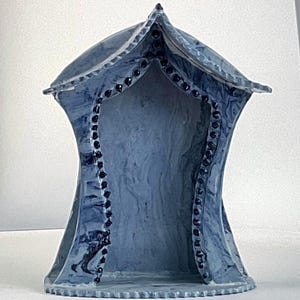 May include: A ceramic structure with a pointed roof and an arched opening, featuring a marbled blue and grey finish. The edges of the arched entrance are decorated with a series of small, raised black dots that follow the curve of the opening.
