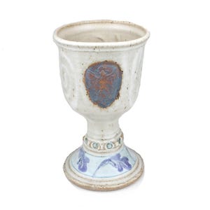 Può includere: Calice in ceramica bianca con un designo blu e marrone. Il calice ha una base larga e una ciotola rotonda.