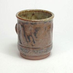 May include: A handmade ceramic mug with a brown and black mottled glaze. The mug features a small handle and a decorative band near the base. The interior has a lighter, greenish-brown hue. This rustic mug is perfect for coffee or tea.