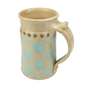 Könnte beinhalten: Eine Keramiktasse mit hellbrauner Glasur und einem geometrischen Muster in Blau und Weiß. Die Tasse hat einen Henkel mit abgerundeter Oberseite.