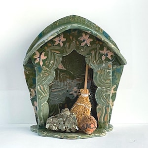 Peut inclure: Une structure en céramique marbrée verte et brune en forme de petite maison avec un toit pointu et des sculptures florales. À l'intérieur se trouvent un balai miniature en paille, un amas de pyrite métallique et un gland brun avec du texte.