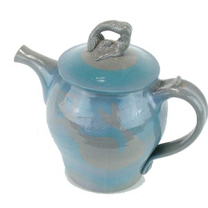 May include: A blue ceramic teapot with a gray glaze and a mermaid-shaped lid. The teapot has a round body and a curved handle.