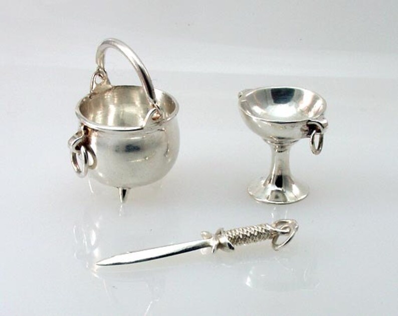 May include: A collection of miniature silver charms, including a cauldron with a handle, a chalice, and a dagger. The cauldron has three small feet. The chalice has a pedestal base and ring handles. The dagger has a detailed handle.