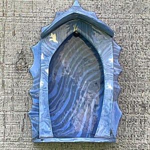 May include: A blue and grey marbled wall-mounted display shelf featuring a pointed gothic arch shape. The structure has a smooth, swirling pattern throughout its surface, with a pointed top and decorative jagged edges along the sides.