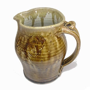 May include: A brown ceramic pitcher with a textured glaze and a raised design of a woman's face. The pitcher has a handle with a unique shape.