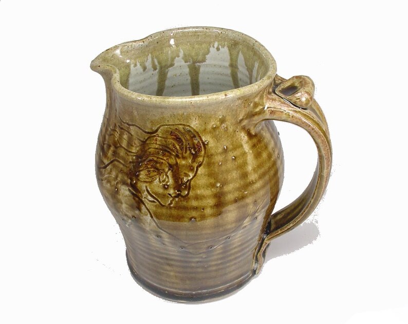 May include: A brown ceramic pitcher with a textured glaze and a raised design of a woman's face. The pitcher has a handle with a unique shape.
