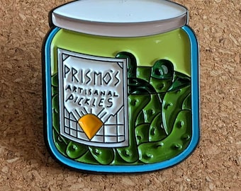 May include: A pin featuring a jar of pickles with the text "Prismos Artisanal Pickles." The jar is green with a blue rim and a white lid. The pickles inside are also green. The pin has a black outline and is set against a corkboard background.