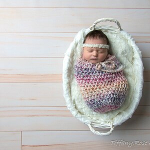 May include: A newborn baby is sleeping in a white wicker basket lined with white faux fur. The baby is wearing a multi-colored crocheted swaddle and a white headband.