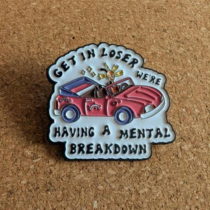 May include: A red convertible car enamel pin with the text "GET IN LOSER WE'RE HAVING A MENTAL BREAKDOWN". The pin features a cartoon-style car with blue wheels and a driver with a shocked expression.