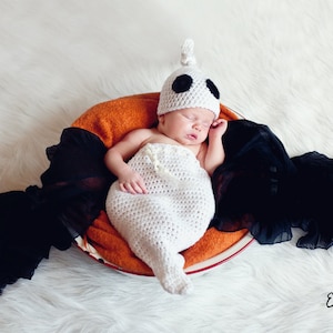May include: A newborn baby wearing a white crocheted ghost costume with black eyes, sleeping in a brown bowl with black fabric draped around it.