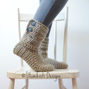 Könnte beinhalten: Ein Paar beige Häkel-Hausschuhe mit schwarzen Knöpfen. Die Schuhe stehen auf einem weißen Holzstuhl.