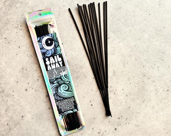 May include: A package of charcoal incense sticks with the text "Sail Away" and a moon and star design. The package is iridescent and contains black incense sticks. A bundle of loose black incense sticks is also present.