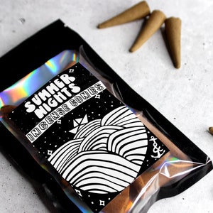May include: A black and white illustrated bag of incense cones with the text "Summer Nights Incense Cones". The bag is made of clear plastic with a black backing and has a holographic iridescent finish. The cones are brown and are scattered around the bag.