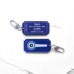 May include: Two blue keyrings with silver clasps. One keyring has the text "TWICE THIS IS FOR WORLD TOUR KIA FORUM LOS ANGELES, CA JANUARY 21-25, 2026". The other keyring has a white and blue graphic.