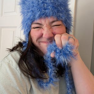 May include: A bright blue fuzzy beanie hat with long, dangling braided ties. The hat is crafted from a textured, shaggy yarn material that creates a soft, voluminous appearance. The design features a rounded crown and two long, matching blue braided tassels.