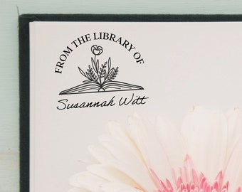 May include: A white book with a black ink stamp that reads "From the Library of Susannah Witt". The stamp features a stylized image of an open book with a flower growing from it. A white gerbera daisy with a pink center is in the background.