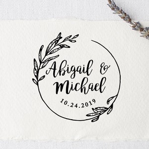 May include: A black and white circular stamp with a floral wreath design. The text inside the wreath reads "Abigail & Michael 10.24.2019".