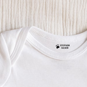 May include: White baby onesie with a black paw print and the name "STEPHEN OLSEN" printed on the neckline.