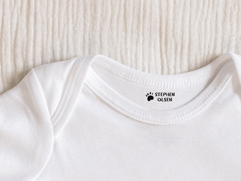 May include: White baby onesie with a black paw print and the name "STEPHEN OLSEN" printed on the neckline.