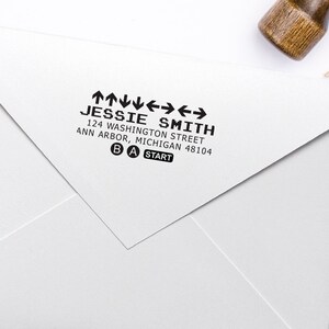 Könnte beinhalten: Ein weißer Umschlag mit einem schwarzen Absenderaufkleber. Der Aufkleber trägt den Text "JESSIE SMITH", "124 WASHINGTON STREET", "ANN ARBOR, MICHIGAN 48104" und eine Grafik eines Videospiel-Controllers mit den Buchstaben "B", "A" und "START" darauf.