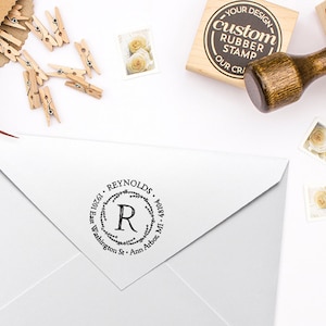 Könnte beinhalten: Ein runder, schwarz-weißer Gummistempel mit dem Text "10261 REYNOLDS" und "R" in einem Kranzdesign. Der Stempel befindet sich auf einem weißen Umschlag.