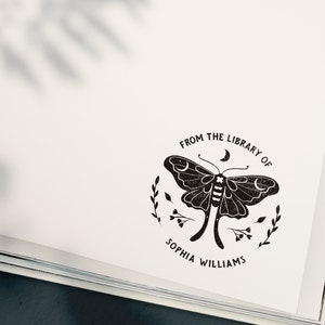 May include: A black and white illustration of a moth with a crescent moon above it. The text "From the Library of Sophia Williams" is written in a circle around the moth.