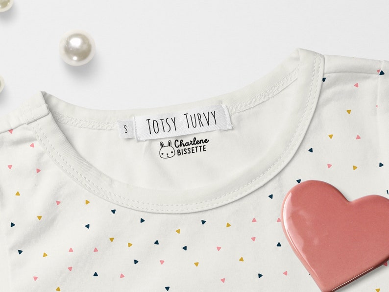 May include: White t-shirt with a pink, gold, and blue triangle pattern. The shirt has a white label with the text "TOTSY TURVY" and a small bunny illustration with the text "Charlene Bissette".