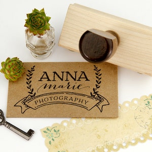 May include: A brown card with the text "ANNA marie PHOTOGRAPHY" in black lettering with a wreath design and a banner. The card is next to a wooden stamp with a black rubber stamp.